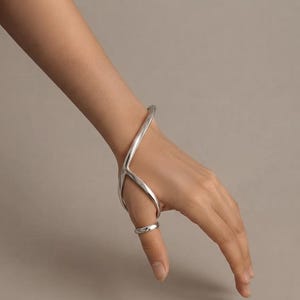 Op de afbeelding: Een gepolijst zilveren handsieraad met een gladde, gebogen metalen band die zich uitstrekt van de pols tot rond de duim. Het sieraad heeft een doorlopend, vloeiend ontwerp met een hoogglans afwerking, waarbij een polsband met een duimring is verbonden.