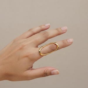 May include: A polished gold-tone ring featuring an elongated, oval-shaped band. The ring has a smooth, continuous metal surface and a wide, open center design that spans across the finger, creating a distinct, minimalist geometric silhouette.