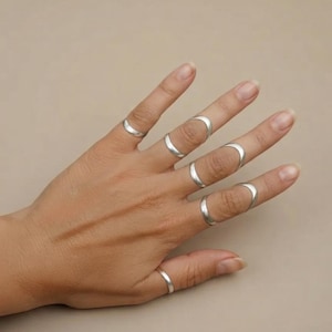 May include: A set of six silver-toned rings worn on the fingers. One ring is placed on the thumb, while the other five rings are positioned on the fingers, with some rings worn at the base and others worn as midi rings between the knuckles.