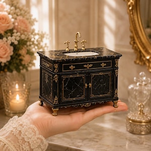 May include: A miniature bathroom vanity featuring a black marble-patterned countertop with gold veining, a white oval sink, and gold-toned faucet fixtures. The cabinet base is black with gold decorative trim, two doors, and ornate gold-colored feet.