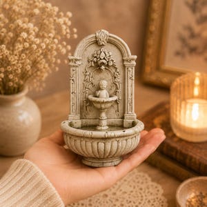 May include: A small, light gray stone-colored fountain ornament featuring a central cherub figure standing in a basin. The structure has an arched back panel with intricate floral carvings, vertical pillars, and a rounded, fluted base bowl.