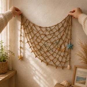 Pode incluir: Uma decoração de parede composta por uma rede de pesca de corda atada em tom bege. A rede é adornada com uma pequena estrela-do-mar texturizada em azul claro no lado direito, uma estrela-do-mar amarela na parte inferior e uma pequena concha.