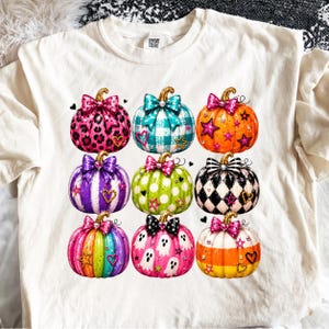 May include: A cream-coloured t-shirt featuring a graphic print of nine pumpkins arranged in a grid. Each pumpkin has a unique pattern, including leopard print, plaid, stripes, polka dots, and ghosts, topped with a decorative bow and star accents.