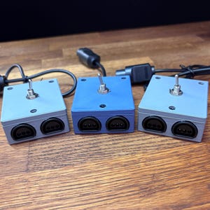 May include: Three rectangular electronic switch boxes with matte finishes in light blue and medium blue. Each unit features a single metal toggle switch on the top surface and two black three-pin ports on the front face, connected to black cables.