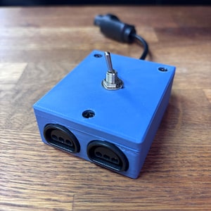 Puede incluir: Una caja de control electrónica azul de forma rectangular que presenta un interruptor de palanca metálico en la parte superior y dos puertos negros en el frente. Un cable negro con conector sale de la parte posterior del dispositivo, asegurado con cuatro tornillos.