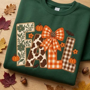 May include: A dark green crewneck sweatshirt featuring the word FALL embroidered in large letters. The F has a green floral pattern, the A has a brown and white cow print, and the Ls feature orange gingham and brown plaid with a large orange bow.