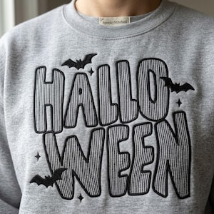 Könnte beinhalten: Ein hellgraues Sweatshirt mit Rundhalsausschnitt und schwarzem Sticktext, der HALLOWEEN in einer fetten, gestreiften Schriftart zeigt. Das Design wird durch kleine schwarze Fledermausstickereien und vierzackige Sterne um die Schrift ergänzt.