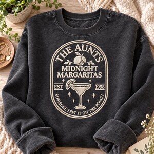 May include: A charcoal gray crewneck sweatshirt featuring an embroidered cream-colored oval emblem. The design includes a lemon, a margarita glass, and the text: THE AUNTS MIDNIGHT MARGARITAS EST 1998 SOMEONE LEFT IT ON THE PORCH with small stars.