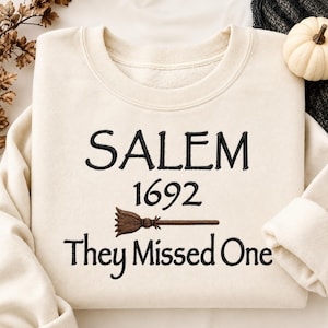 以下が含まれることがあります： クリーム色のクルーネックトレーナー。黒い刺繍で「SALEM 1692」および「They Missed One」という文字が施されており、年号と最後の行の間には茶色の刺繍で小さなほうきのアイコンが配置されています。