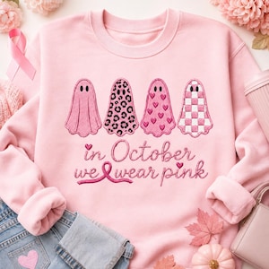 May include: A light pink crewneck sweatshirt featuring four ghost illustrations with different patterns: solid pink, leopard print, heart print, and a checkered pattern. Below the ghosts, the text reads: in October we wear pink.