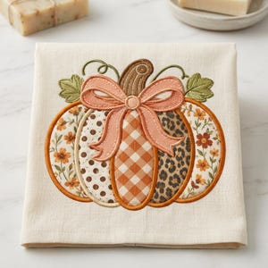 May include: A cream-colored fabric towel featuring an embroidered pumpkin design. The pumpkin is composed of five vertical segments with distinct patterns: floral, polka dot, gingham, leopard print, and floral. A peach-colored bow sits on top.