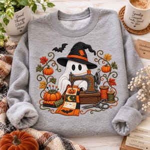 Pode incluir: Uma camisola cinzenta mesclada de gola redonda com um bordado de um fantasma branco com um chapéu de bruxa preto a usar uma máquina de costura. O design inclui abóboras cor de laranja, folhas de outono, carretéis de linha e tesouras.