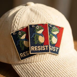 May include: A cream-colored corduroy baseball cap featuring three rectangular embroidered patches attached with safety pins. Each patch displays a green frog wearing a blue scarf against a red and dark blue background with the text RESIST.