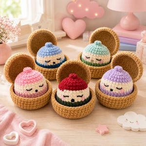 May include: Five small, round, crocheted figures with closed eyes and rosy cheeks, each nestled inside an open, tan, textured crocheted shell. The figures feature colorful hats in blue, mint green, pink, red, and purple, with cream-colored faces.