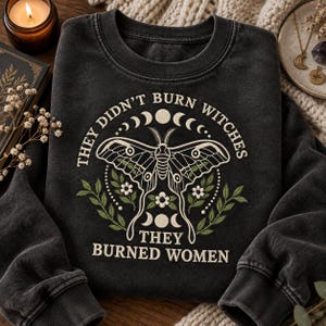 Può includere: Una felpa girocollo grigio antracite con un ricamo raffigurante una falena, fasi lunari e rametti floreali. Il testo recita: "THEY DIDN'T BURN WITCHES THEY BURNED WOMEN." Il disegno utilizza fili color crema e verde oliva sul tessuto scuro.