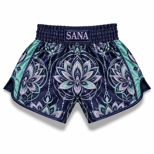 May include: Navy blue athletic shorts featuring a repeating lotus flower pattern in shades of light purple and teal. The waistband is elasticised with the text SANA centred on a rectangular patch. The sides have teal panels.