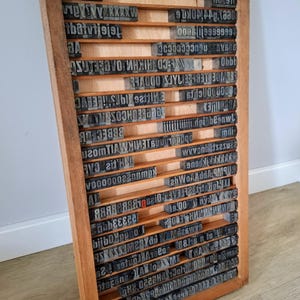 Op de afbeelding: Een rechthoekige houten letterbak met meerdere horizontale vakken gevuld met diverse metalen en houten letterblokken. Het frame is gemaakt van lichtbruin hout en de binnenzijde bevat rijen met uiteenlopende typografische karakters.