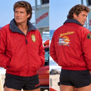 May include: A red zip-up jacket with a dark blue collar, featuring a Baywatch logo on the chest and back, paired with black shorts. The back graphic displays a lifeguard tower and a sunset. The jacket has patches on the sleeves and ribbed cuffs.