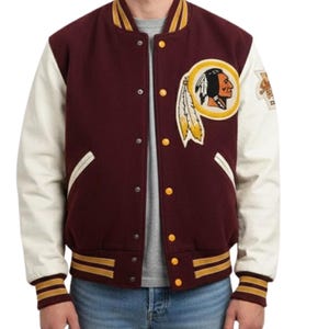 Pode incluir: Uma jaqueta varsity bordô com mangas cor de creme, apresentando um emblema circular no peito esquerdo com o perfil de uma figura nativa americana com penas. A jaqueta tem punhos, gola e bainha canelados com listras amarelas e bordô.