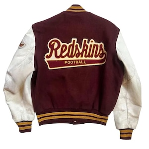 May include: A burgundy varsity jacket with white sleeves and striped ribbed cuffs and hem. The back features a large embroidered patch with the text Redskins FOOTBALL in burgundy and gold. A small logo patch is visible on the right sleeve.