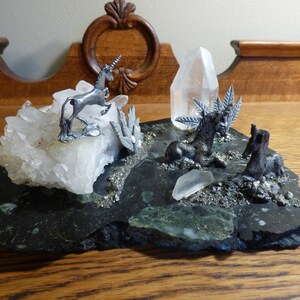 May include: A silver unicorn figurine standing on a bed of white quartz crystals, surrounded by two silver unicorn foals on a black slate base with green and gold accents.