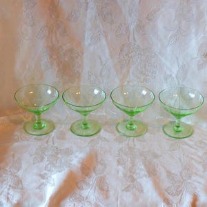 May include: Four green glass dessert cups with a delicate etched design. The cups are arranged in a row on a white tablecloth with a floral pattern.