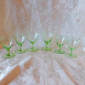 May include: Six green glass drinking glasses with a fluted design. The glasses are arranged in a row on a white tablecloth.