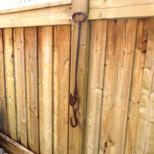 Peut inclure: Un crochet en métal rouillé avec une poignée, suspendu à une clôture en bois. Le crochet a une longue tige et deux crochets incurvés. La clôture est faite de planches de bois verticales.