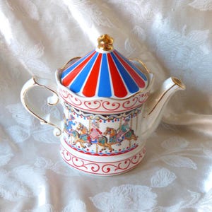 May include: A ceramic teapot with a carousel design. The lid is striped red and blue, topped with a gold finial. The body features a carousel scene with riders on horses. Gold trim accents the handle and spout.