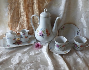 May include: A white porcelain tea set with floral designs. The set includes a teapot, creamer, sugar bowl, two teacups, and two saucers. The set is on a white tablecloth with a lace overlay.
