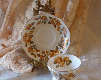 May include: A white teacup and saucer with a gold trim and a floral pattern of yellow roses. The teacup and saucer are on a gold stand.