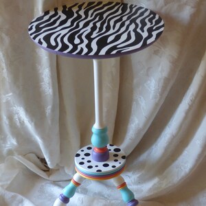 May include: A small, white, zebra-print table with a colourful base. The base has a black and white polka dot design, a turquoise and orange band, and three legs with purple and orange accents.