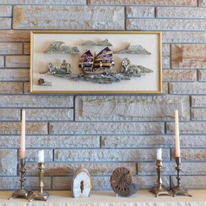May include: A framed picture of a boat with purple and gold sails, set against a backdrop of clouds and water. Two brass candle holders flank a geode and a fossil. The artwork and decor are set against a stone brick wall.