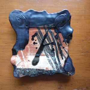 May include: A small, square ceramic dish with a black rim and an abstract design in shades of brown, black, and white.