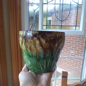 Peut inclure: Un cache-pot en céramique doté d'un vernis brillant passant d'un brun foncé profond au niveau du bord à un vert herbe éclatant à la base. La partie inférieure est ornée d'un motif en relief représentant un soleil avec des rayons.