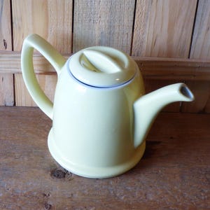 May include: A pale yellow ceramic teapot with a curved handle and spout. The lid has a small handle and a thin blue line around the edge. The teapot is sitting on a wooden surface.