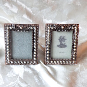 May include: Two rectangular bronze-coloured picture frames with pearl and rhinestone accents. Each frame has a small, rectangular opening for a photo. One frame contains a grey background, and the other has a silhouette image.