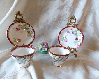 May include: Two vintage teacups and saucers with a floral design of tulips and daffodils. The cups and saucers have a red and gold trim. The cups and saucers are on a decorative stand.