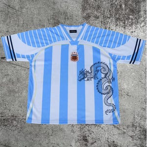 May include: A short-sleeve jersey featuring vertical light blue and white stripes. The chest displays a centered AFA emblem with three stars above it. A large, detailed black line-art dragon graphic is positioned on the lower right side.
