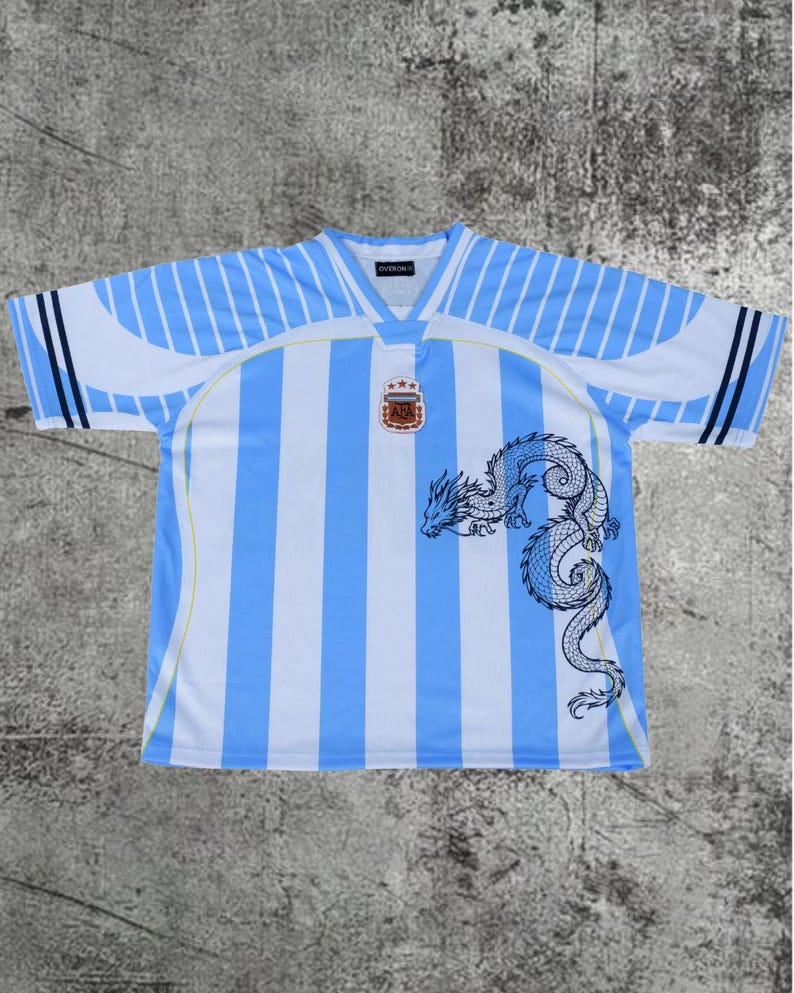 May include: A short-sleeve jersey featuring vertical light blue and white stripes. The chest displays a centered AFA emblem with three stars above it. A large, detailed black line-art dragon graphic is positioned on the lower right side.