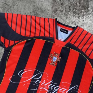 May include: A short-sleeved sports jersey featuring vertical black and red stripes. The chest displays the Portugal national team crest and the word Portugal in white cursive script. The collar is black and red with an Overon brand label inside.