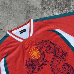 May include: A red jersey with a white and dark green V-neck collar. The shirt features a dark blue dragon graphic and a yellow and red crest with the text 1909. A black label inside the collar displays the text OVERON. The fabric has a subtle pattern.