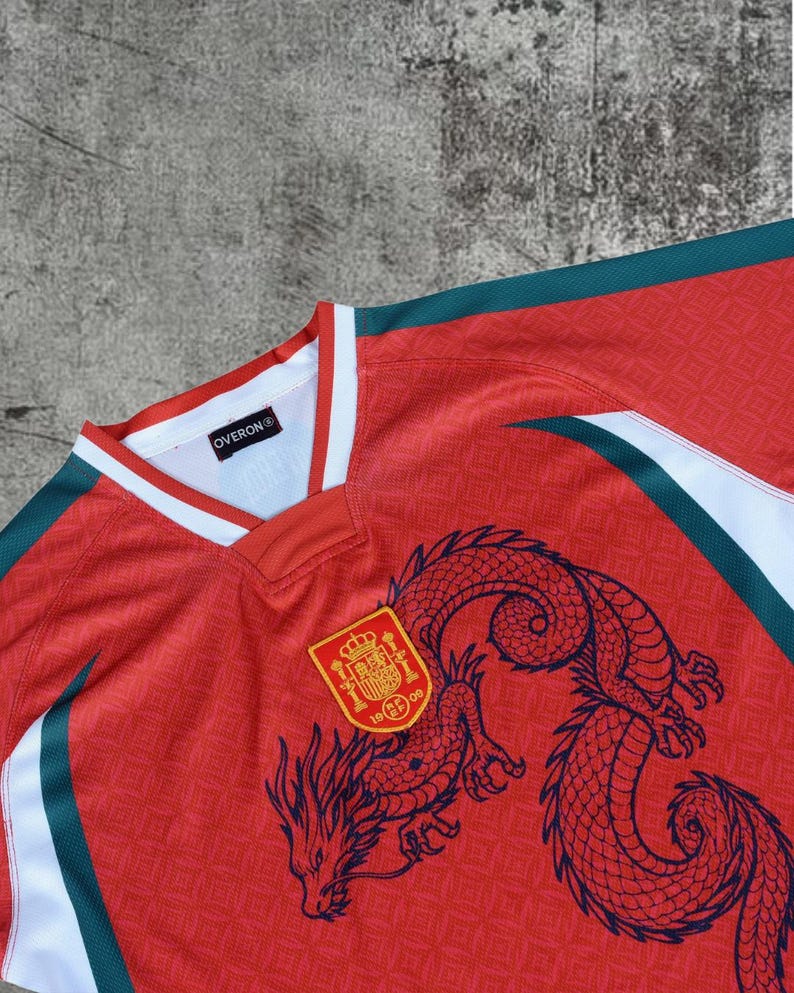 May include: A red jersey with a white and dark green V-neck collar. The shirt features a dark blue dragon graphic and a yellow and red crest with the text 1909. A black label inside the collar displays the text OVERON. The fabric has a subtle pattern.