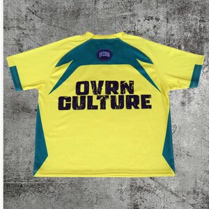 May include: A short-sleeve t-shirt in bright yellow with dark teal accents on the shoulders and sides. The back features a dark purple circular logo with the text OVERON and the large, distressed dark purple text OVRN CULTURE printed below.