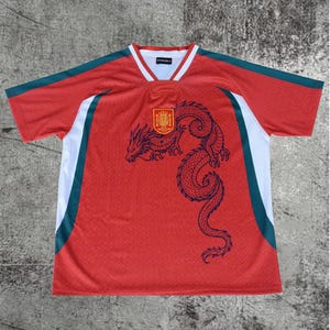 May include: A red short-sleeve jersey featuring a dark blue dragon graphic on the front, a yellow and red crest emblem, and white and dark green side panels. The collar is white with red trim, and the interior label displays the text OVERON.