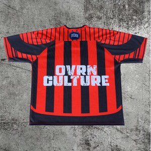 May include: A short-sleeved jersey featuring vertical red and black stripes. The center of the chest displays the white text OVRN CULTURE in a distressed font. A small blue circular patch with the word OVERON is located at the neckline.