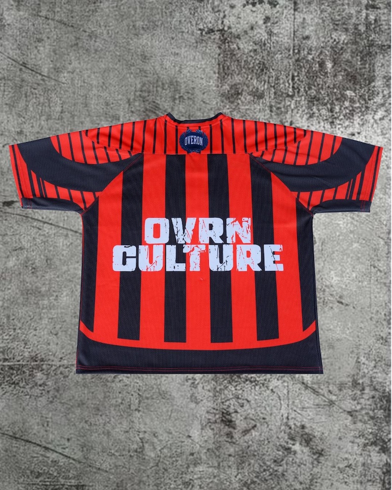 May include: A short-sleeved jersey featuring vertical red and black stripes. The center of the chest displays the white text OVRN CULTURE in a distressed font. A small blue circular patch with the word OVERON is located at the neckline.