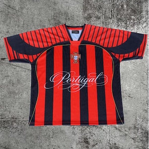 May include: A short-sleeve jersey featuring vertical red and black stripes. The chest displays a crest and the word Portugal in white cursive script. The V-neck collar is black, and the shoulders have black curved panels with thin yellow piping.