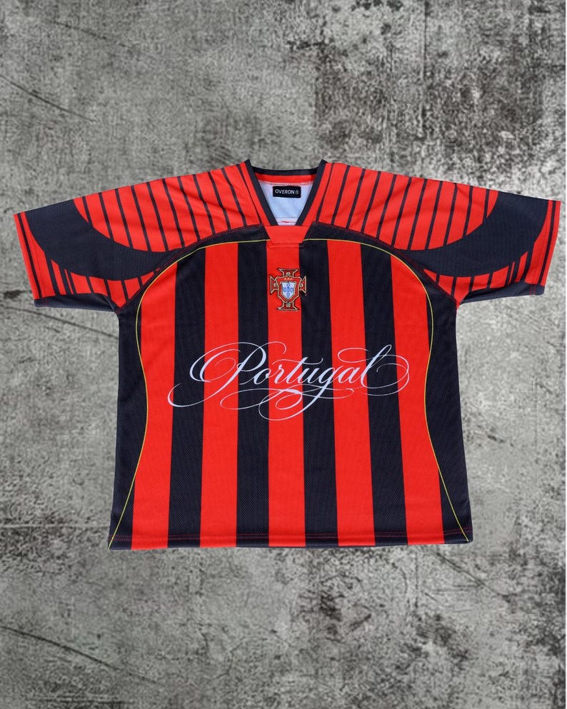 May include: A short-sleeve jersey featuring vertical red and black stripes. The chest displays a crest and the word Portugal in white cursive script. The V-neck collar is black, and the shoulders have black curved panels with thin yellow piping.