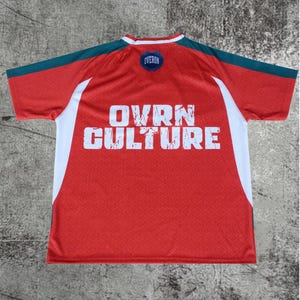 May include: A red short-sleeved jersey with dark teal shoulder panels and white side accents. The back features the white text OVRN CULTURE in a distressed font and a small circular blue patch at the neckline with the text OVERON.